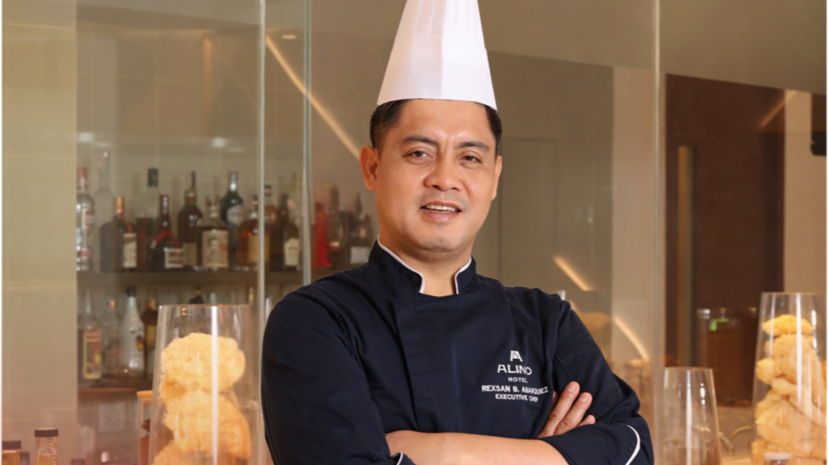 Chef in a white hat and black uniform stands with arms crossed in a restaurant kitchen.