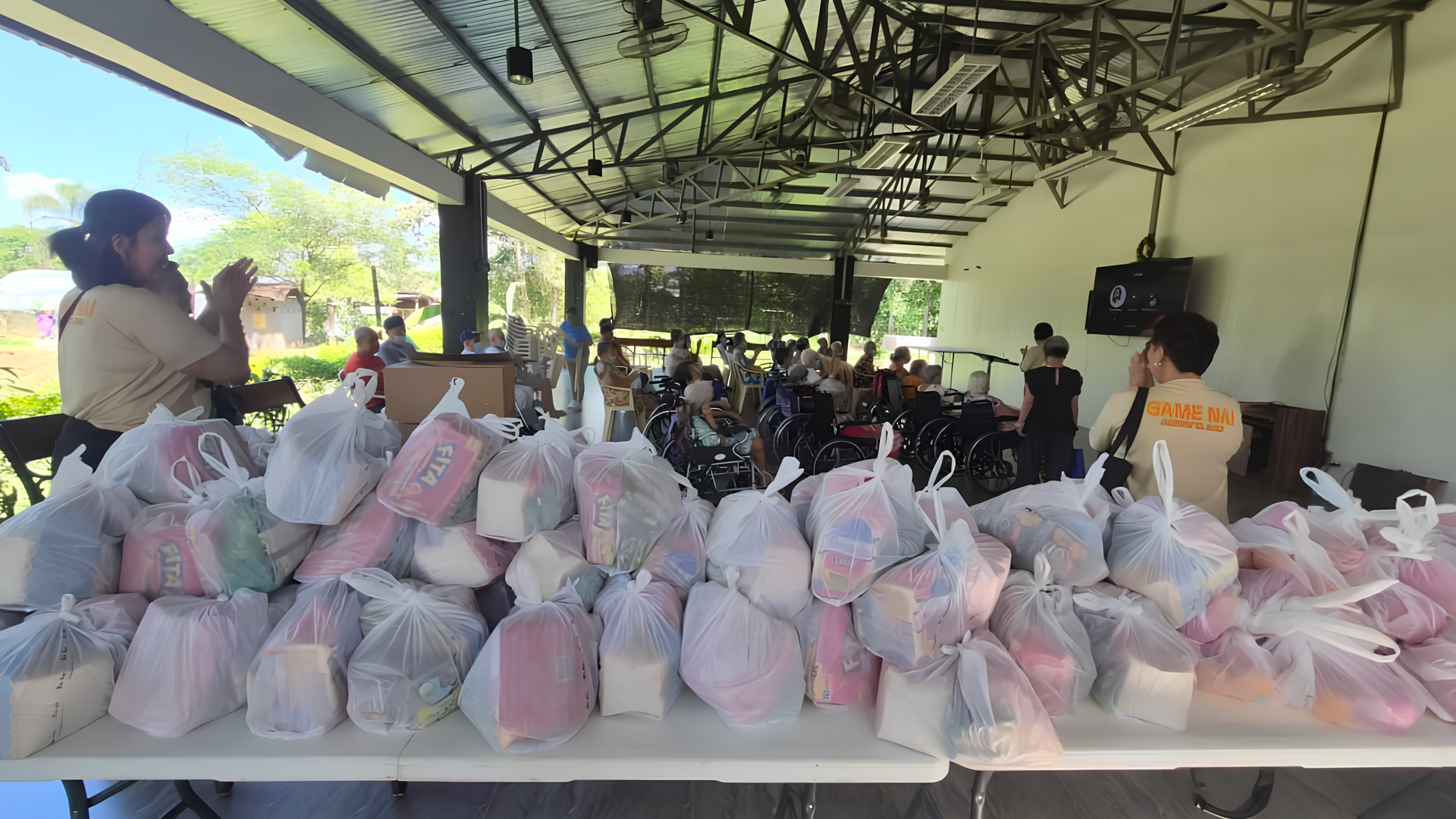 People receiving aid under a covered pavilion, with tables full of bagged supplies in the foreground