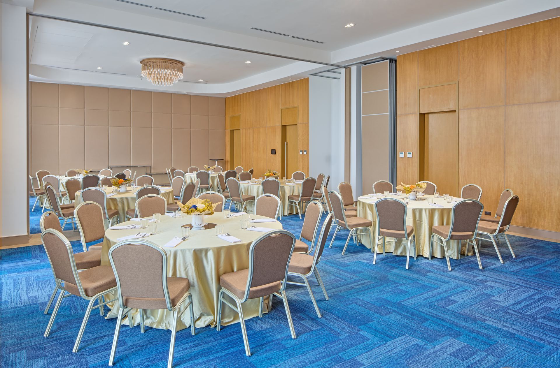 Banquet hall with round tables covered in beige tablecloths, surrounded by brown chairs on a blue patterned carpet.