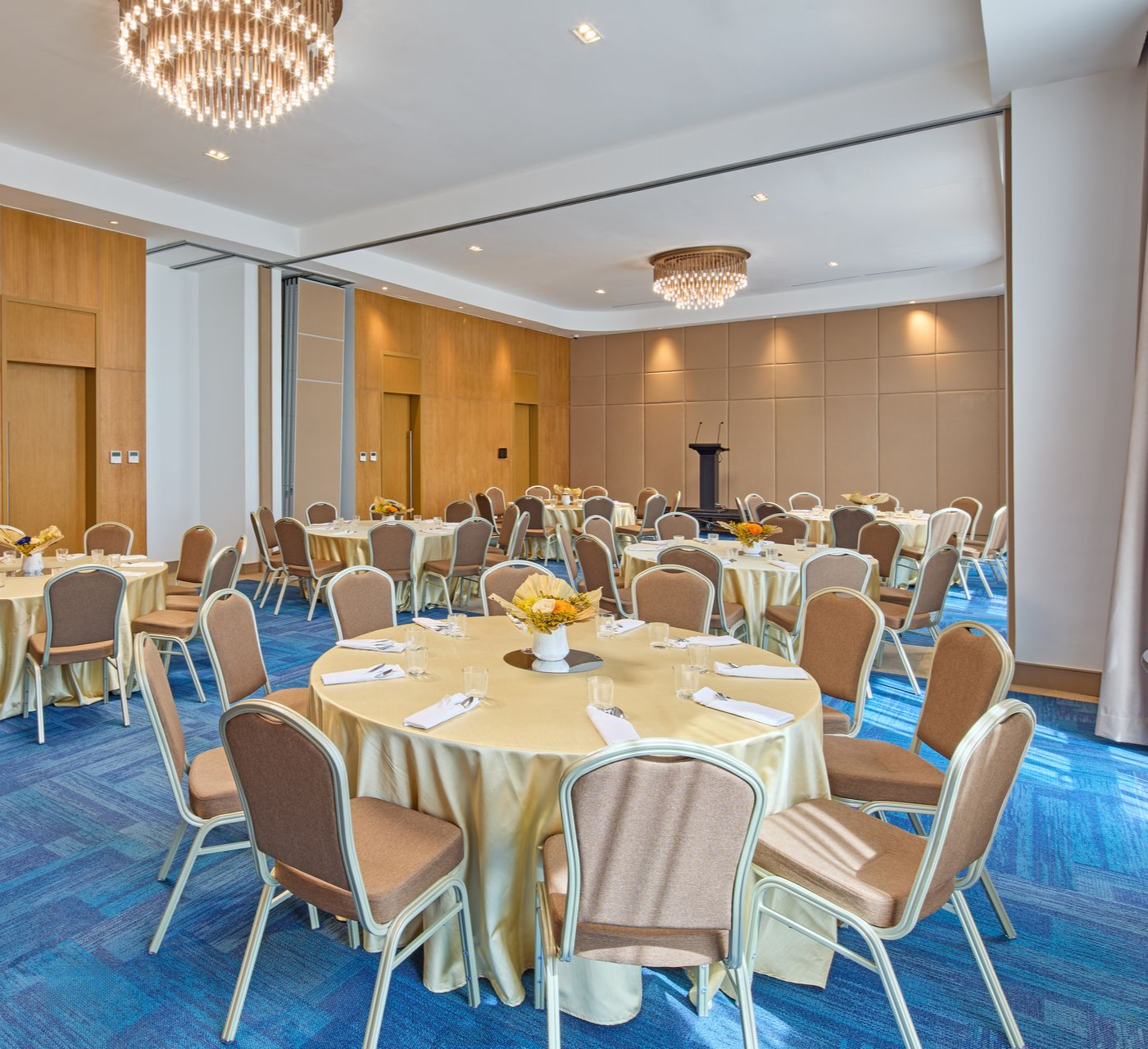 A conference room set up for a banquet with round tables, gold tablecloths, tan chairs, and blue patterned carpet.