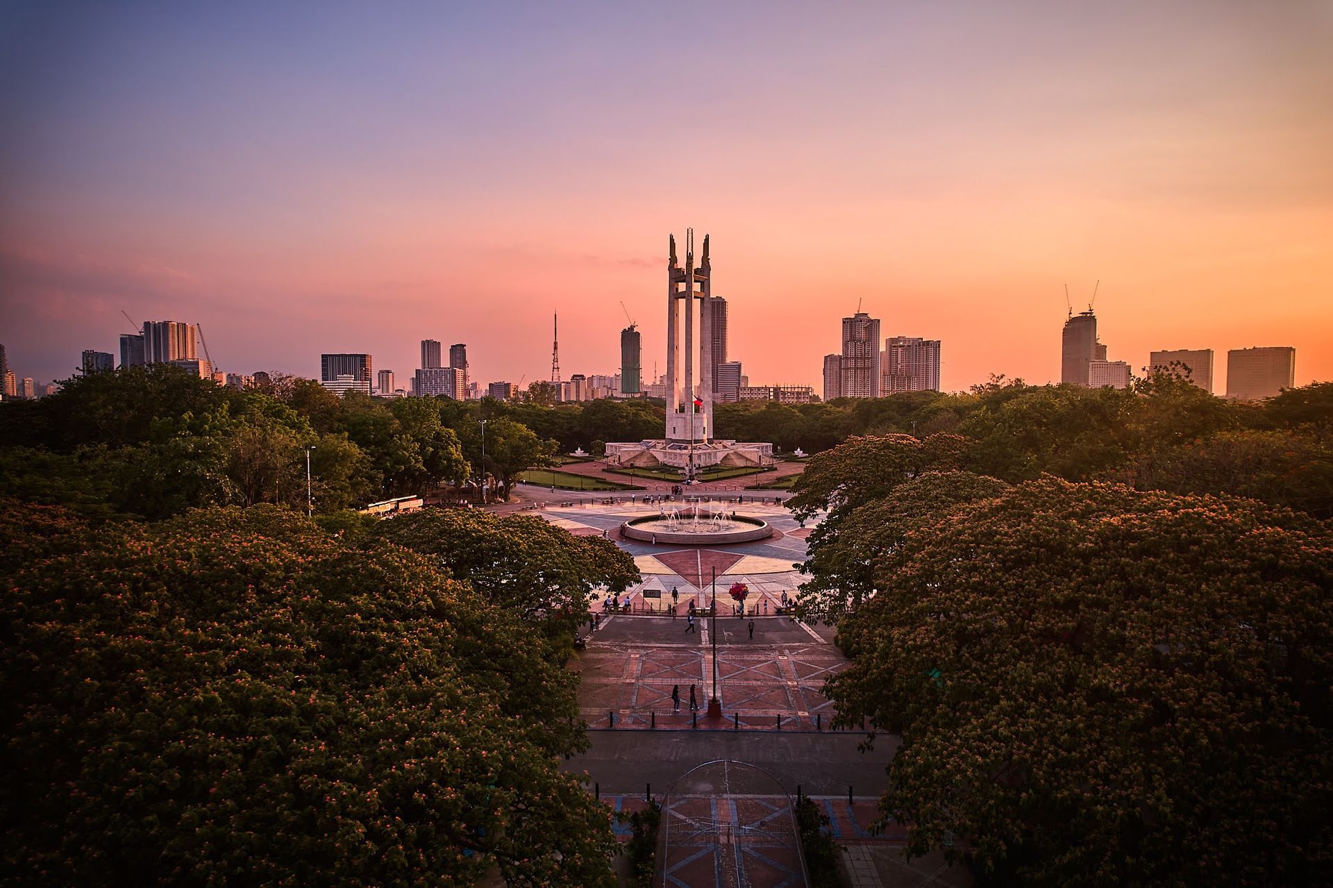 Sunset over a city skyline framed by trees and a wide walkway with fountains below