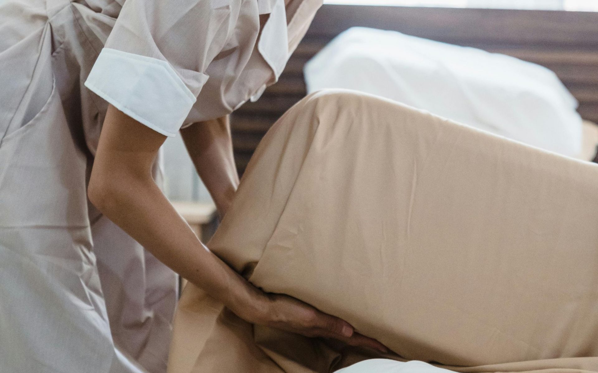 Person making a bed, adjusting a tan sheet in a room.