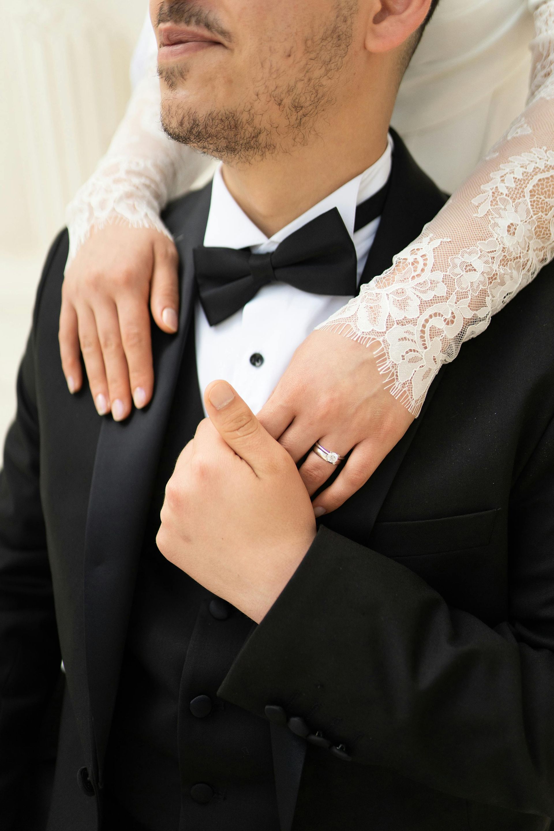 A person in a black tuxedo with a bowtie, with another person's lace-sleeved arms embracing them and an engagement ring.