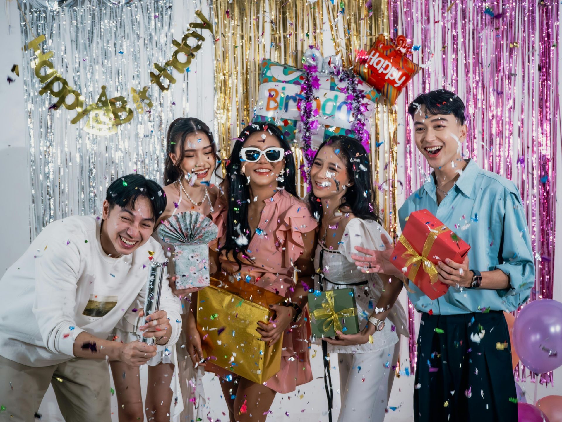 Four friends smiling and holding gifts amid falling confetti in front of a colorful streamer birthday backdrop.