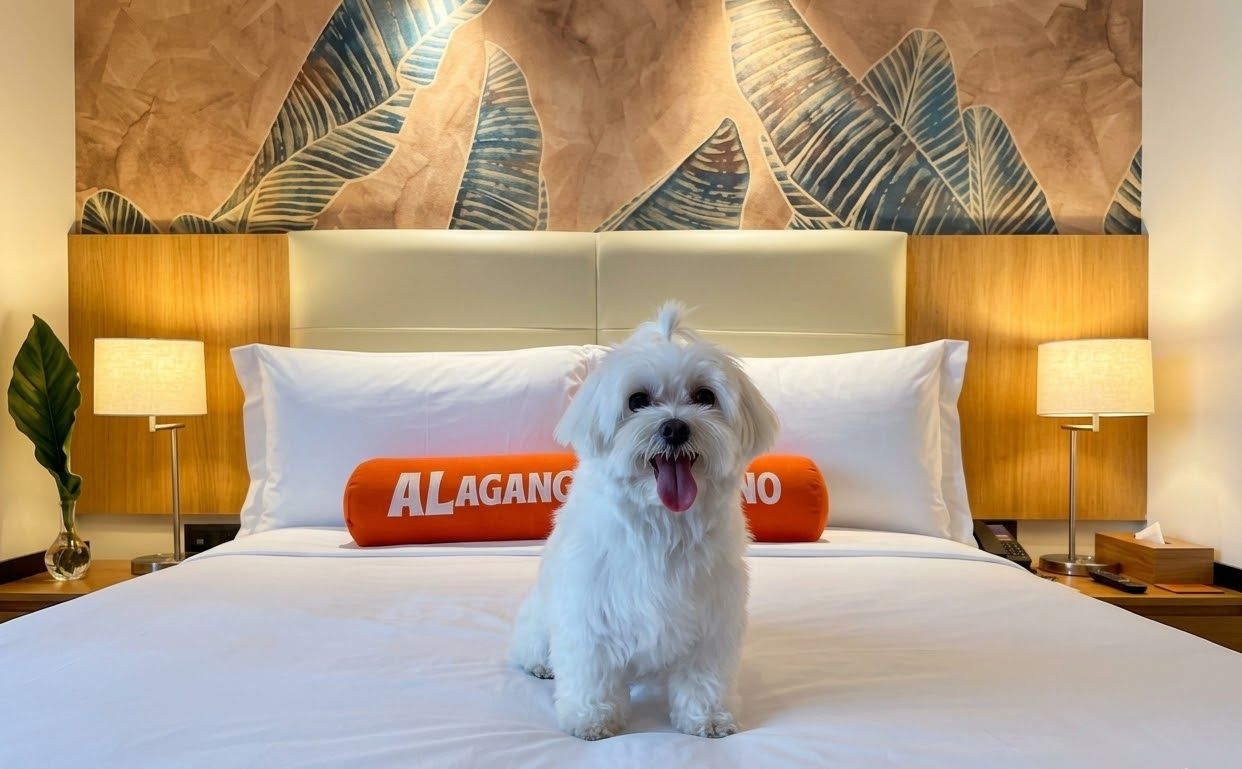 White dog with tongue out sits on a hotel bed, orange pillow, lamps, leaf decor in the background.