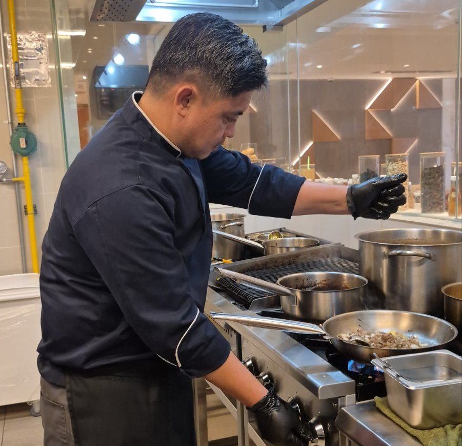 Chef in black uniform cooking at a stainless steel stovetop in a commercial kitchen.