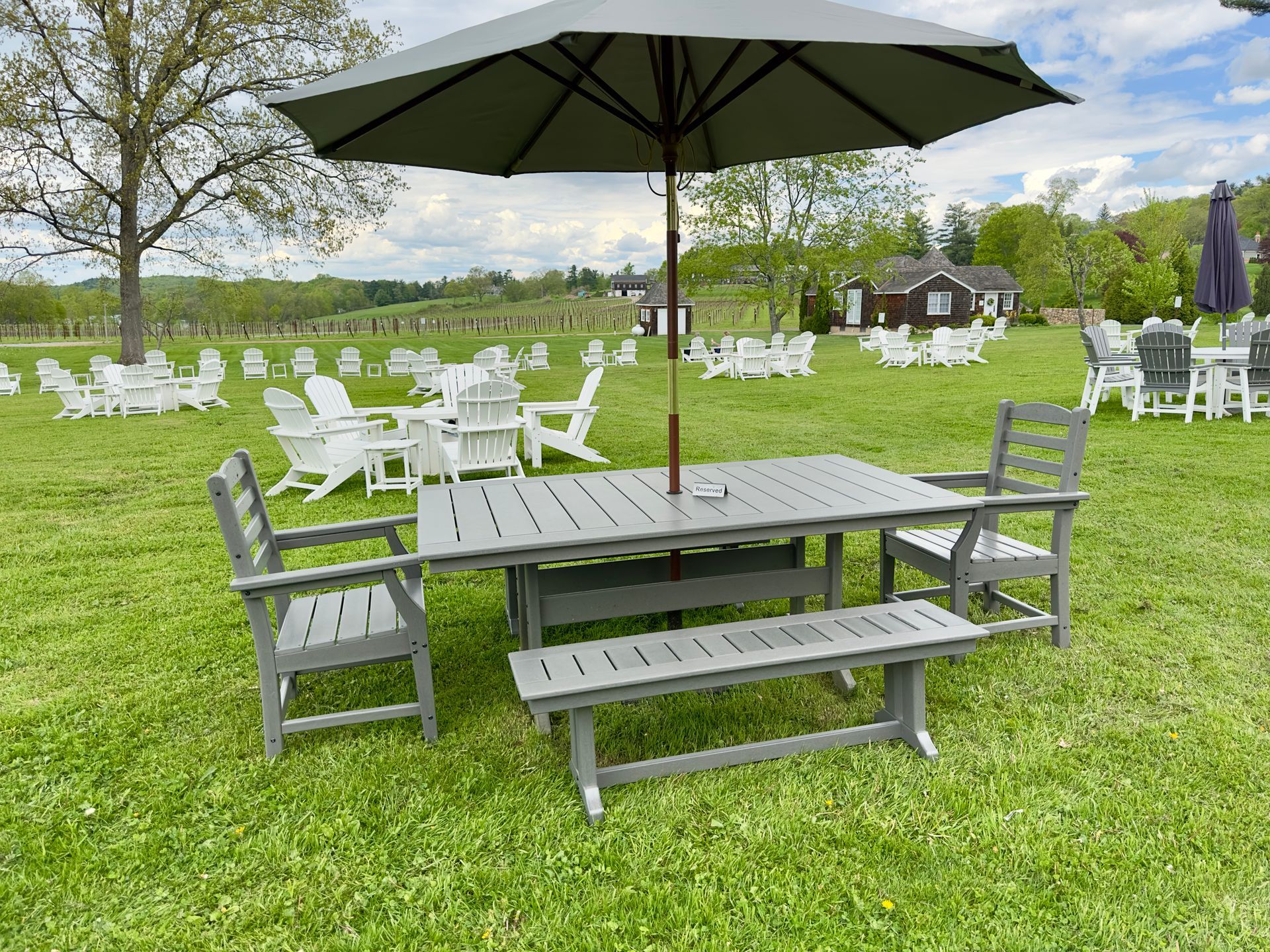 Outdoor picnic table with benches and a black umbrella on a grassy lawn surrounded by white chairs and trees