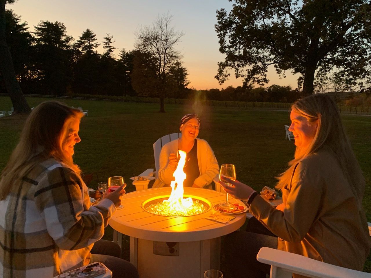 Three people sit around a glowing fire pit outdoors at sunset, holding drinks and talking.