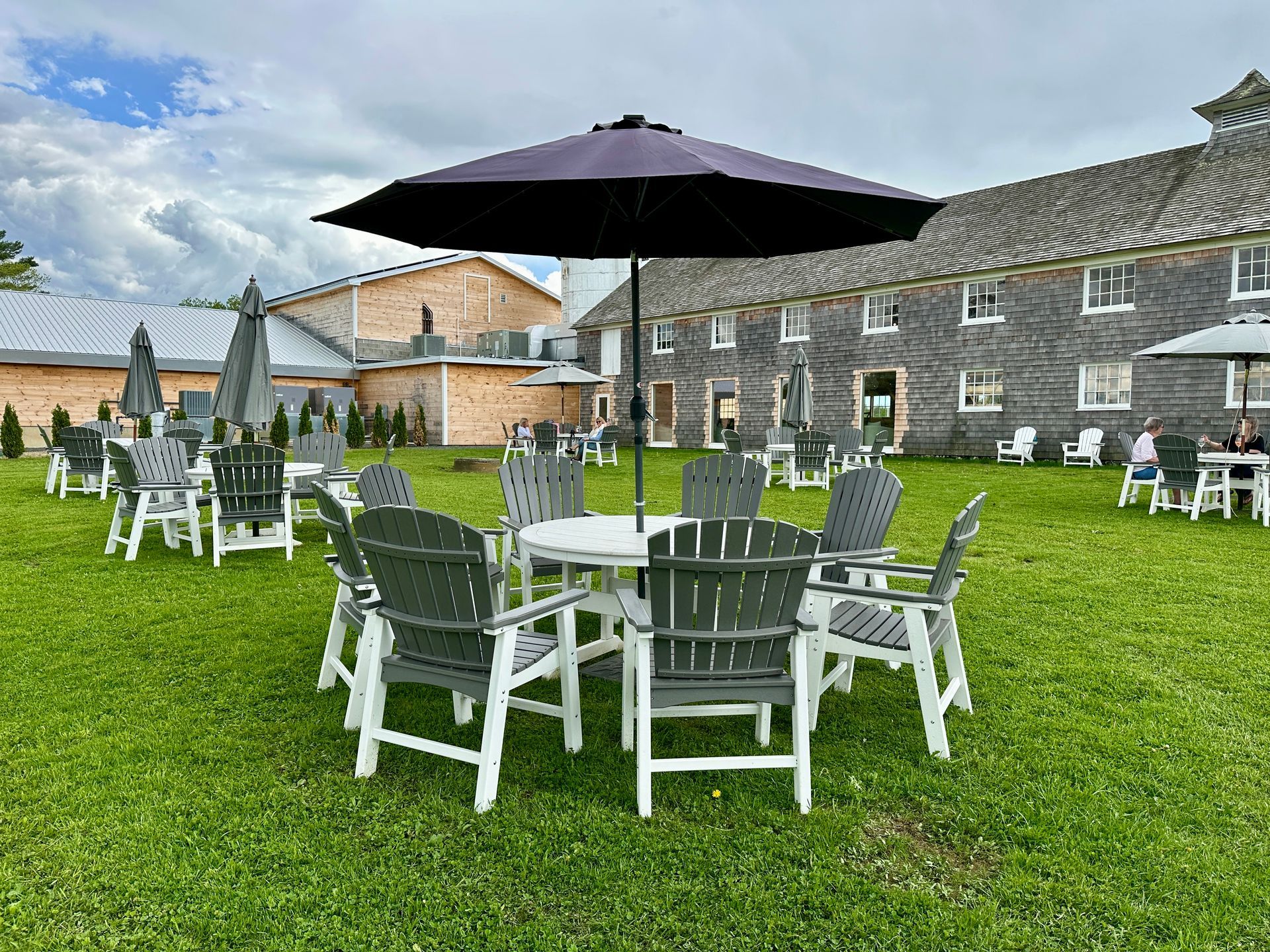 Outdoor patio with white chairs and tables on a grassy lawn beside stone buildings, under a large black umbrella