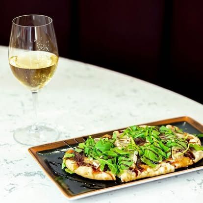 Wine glass beside a plated flatbread topped with greens on a marble table