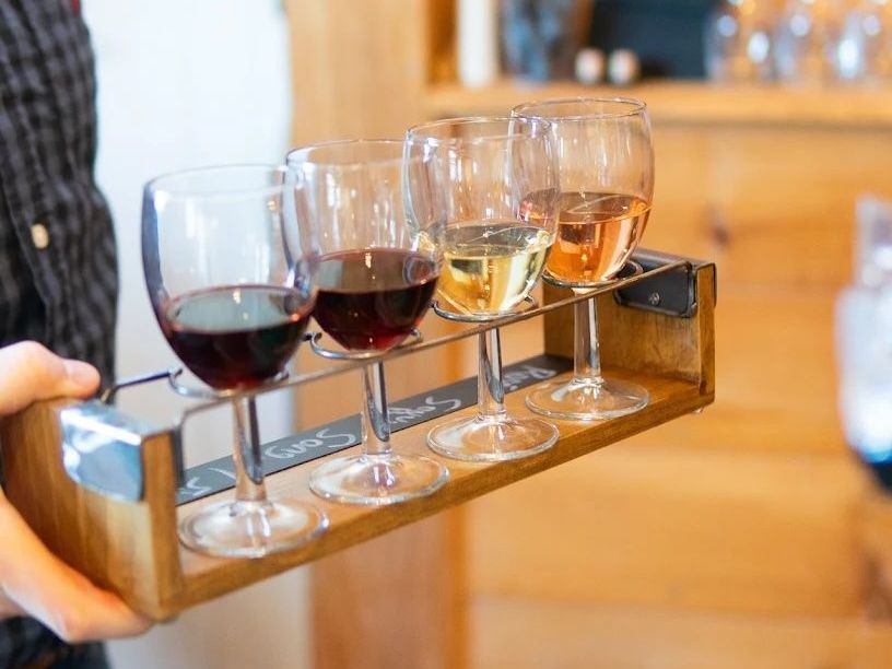 Wine tasting flight: Four glasses with red, white, and rosé wines on a wooden tray held by a person.