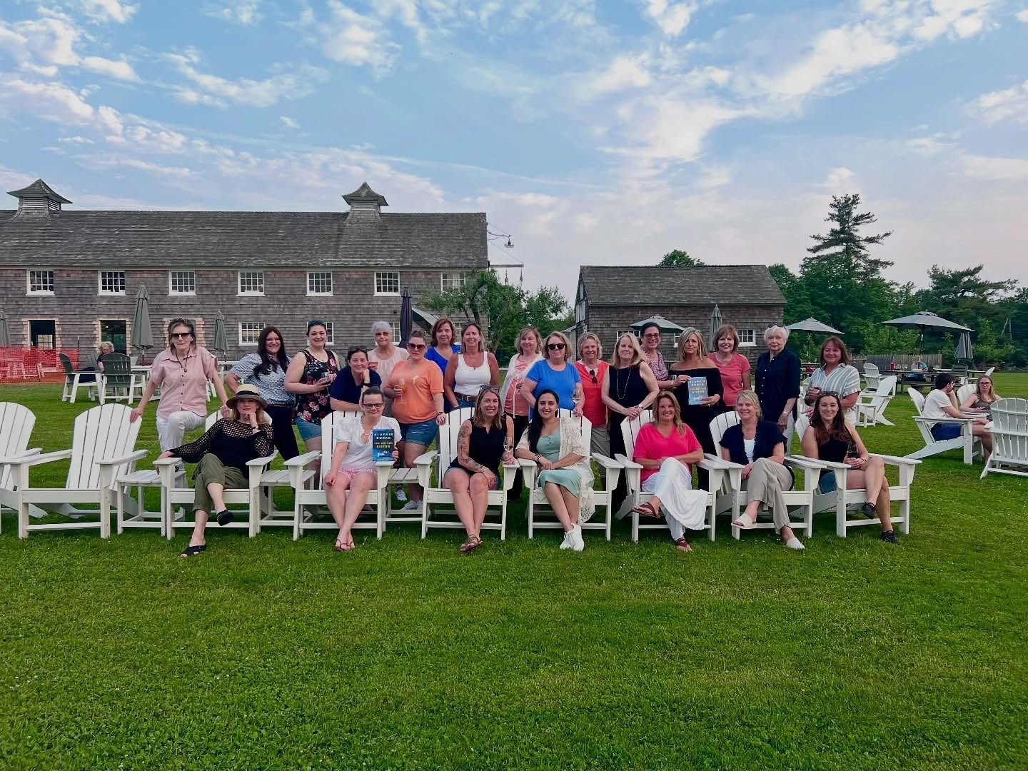 Large group seated on white chairs outdoors on a lawn in front of a stone building, smiling at the camera.