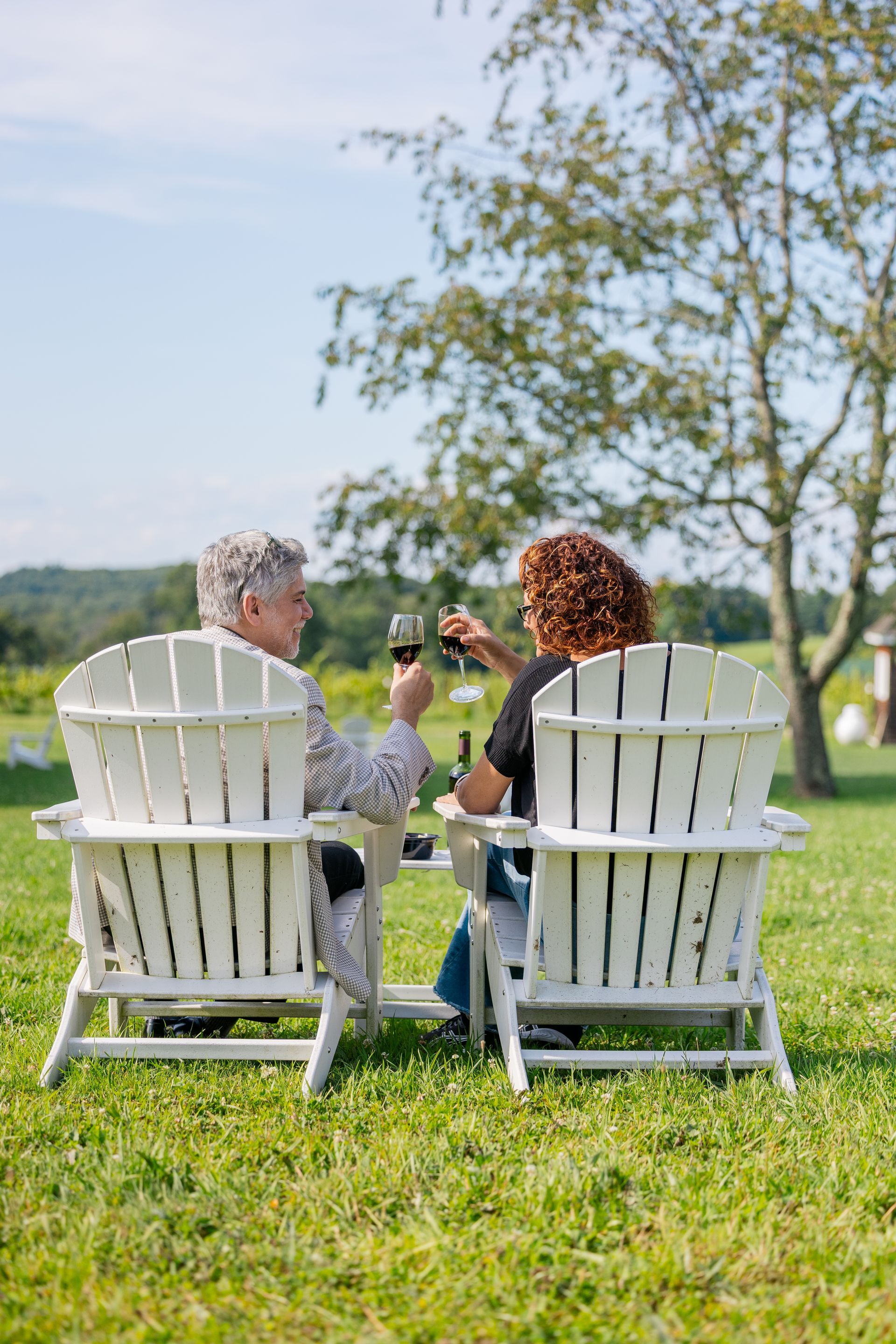 Two people sit in white Adirondack chairs on a grassy lawn, toasting glasses beside a lake.