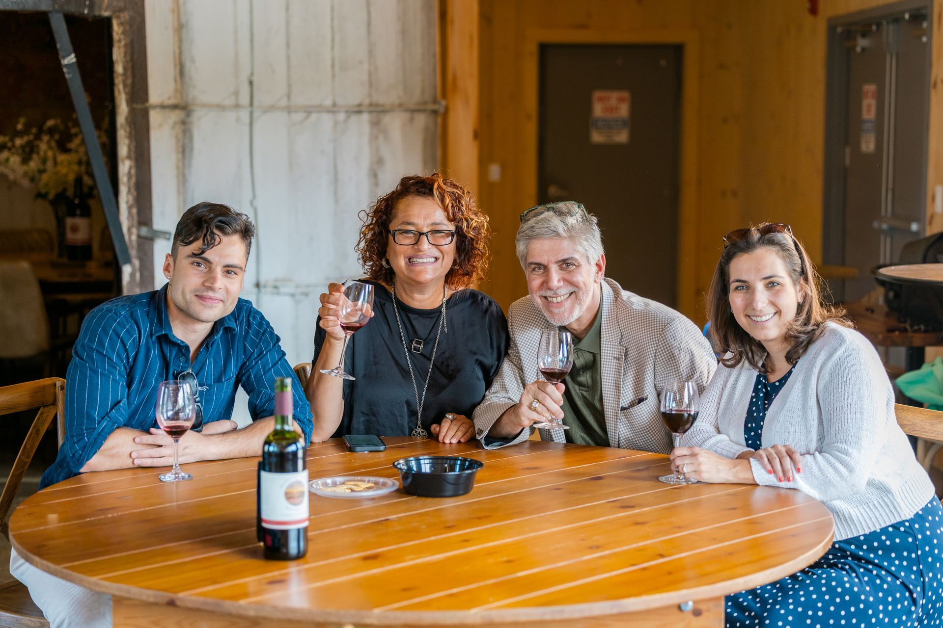 Four people seated around a wooden table in a rustic room, smiling and holding drinks.