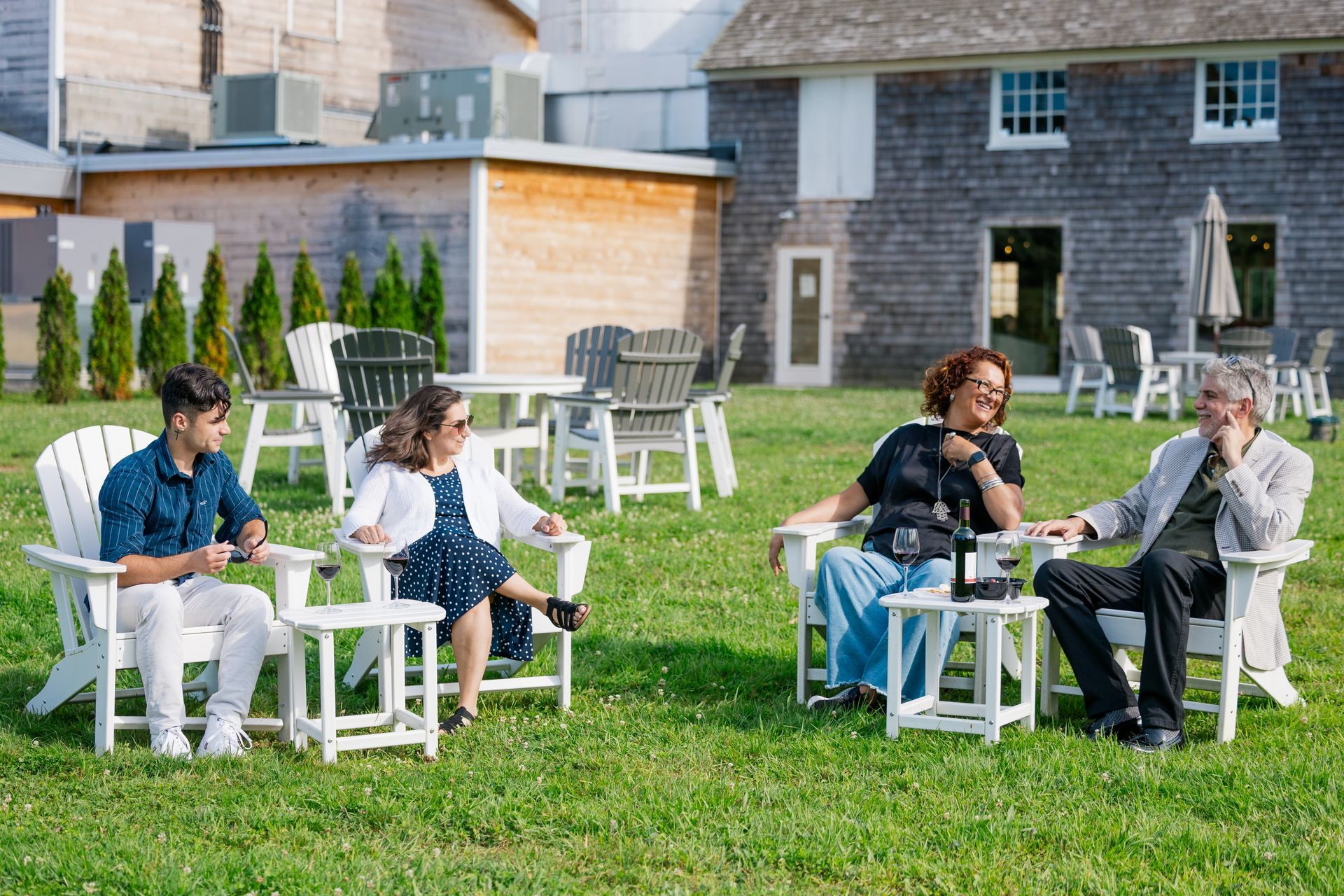 People sitting outdoors on white chairs around small tables on a grassy lawn near houses.