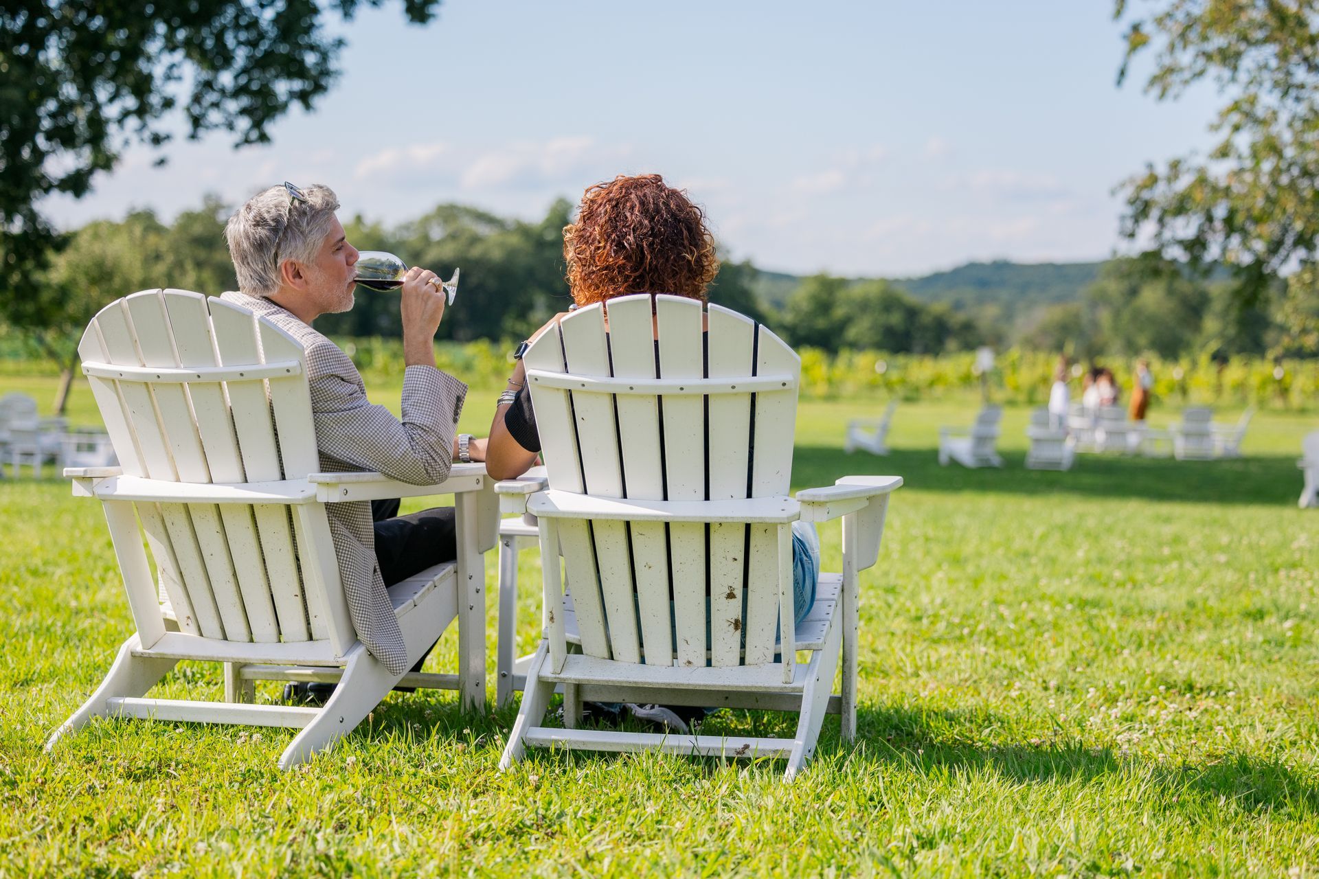 Two people in white Adirondack chairs talking on a sunny lawn with a pond and hills in the background