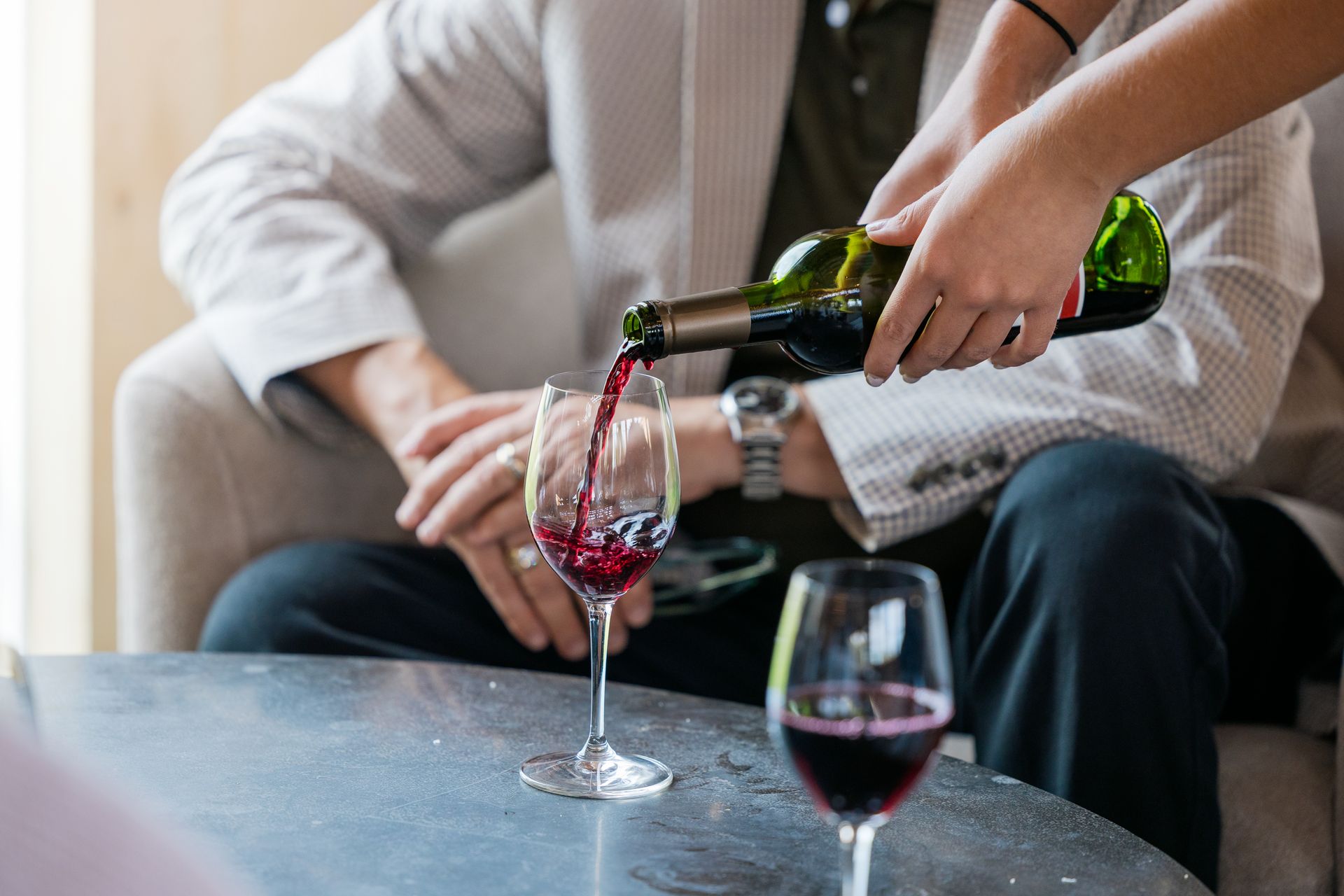 Person pouring red wine into a glass at a table while another sits nearby