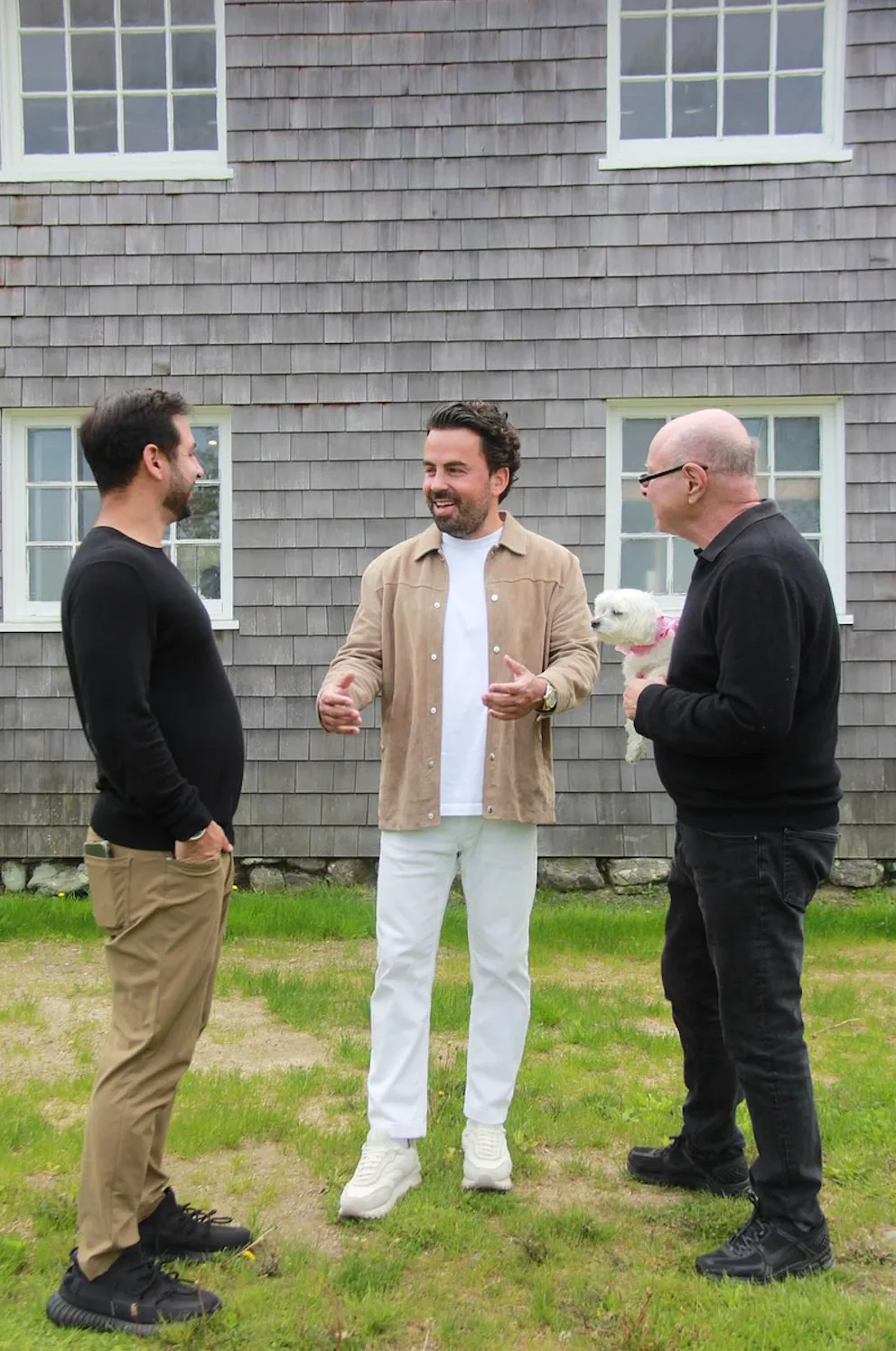 Three men chatting outdoors in front of a gray-shingled house, one holding a drink
