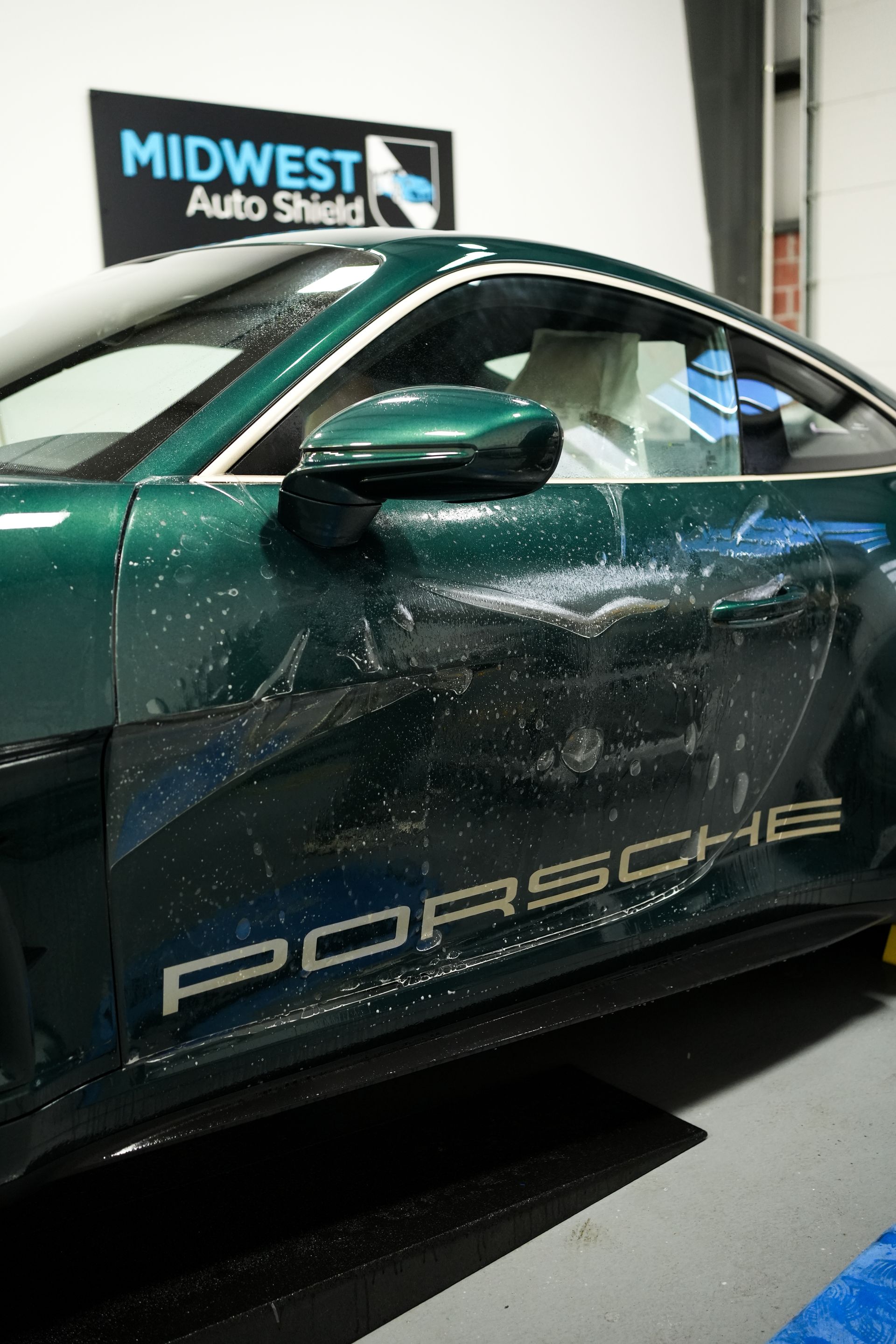 Green Porsche sports car in a shop with a visible “Midwest Auto…” sign in the background