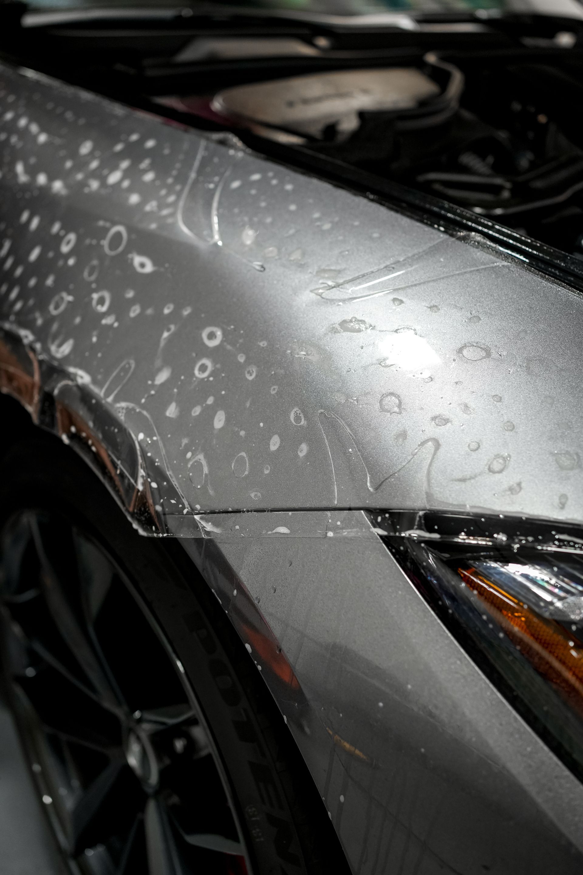 Close-up of a silver car hood and headlight covered in water droplets.