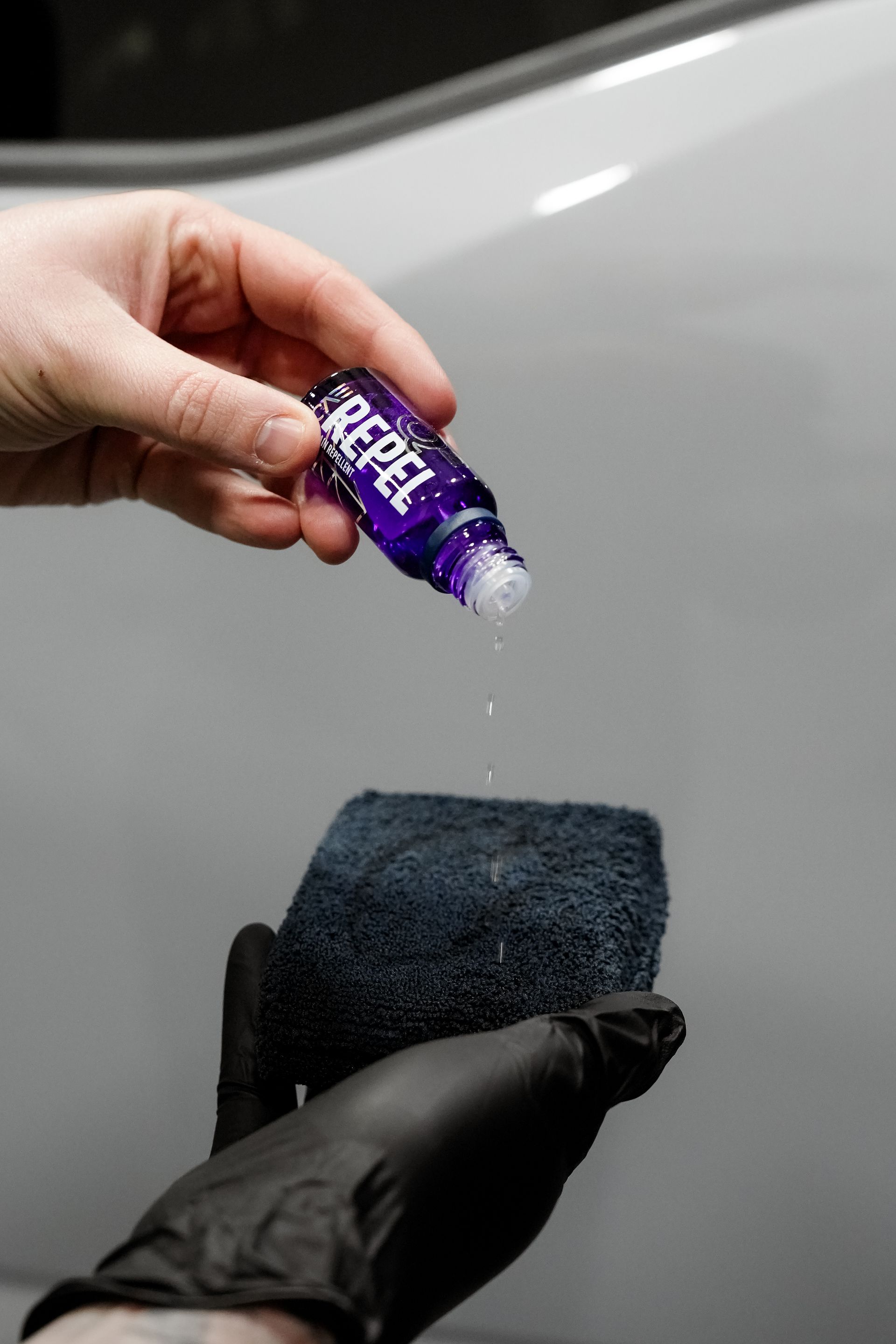 Hand pouring purple liquid onto a black applicator pad over a car surface