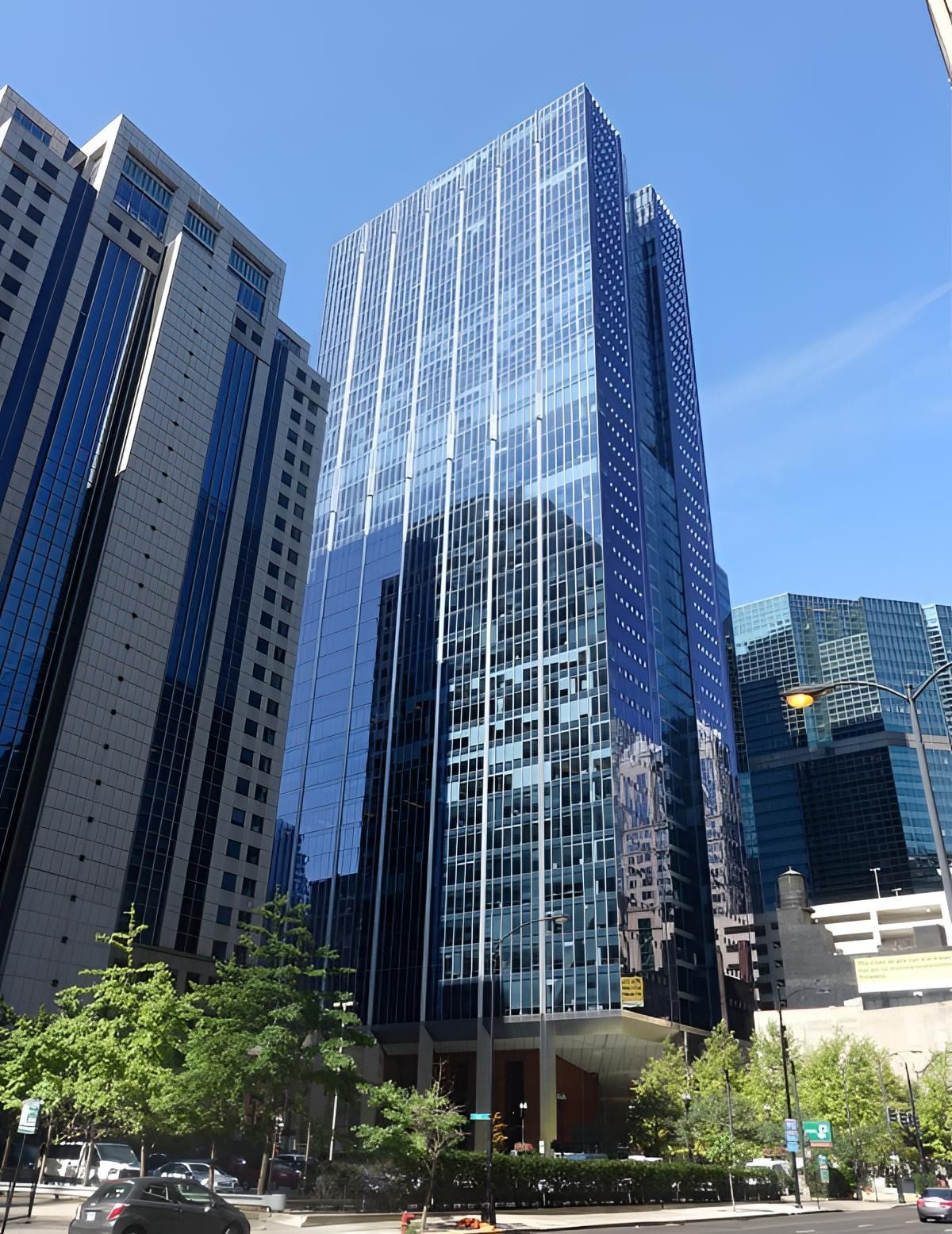Tall glass skyscraper reflecting blue sky, surrounded by neighboring high-rise buildings on a city street.