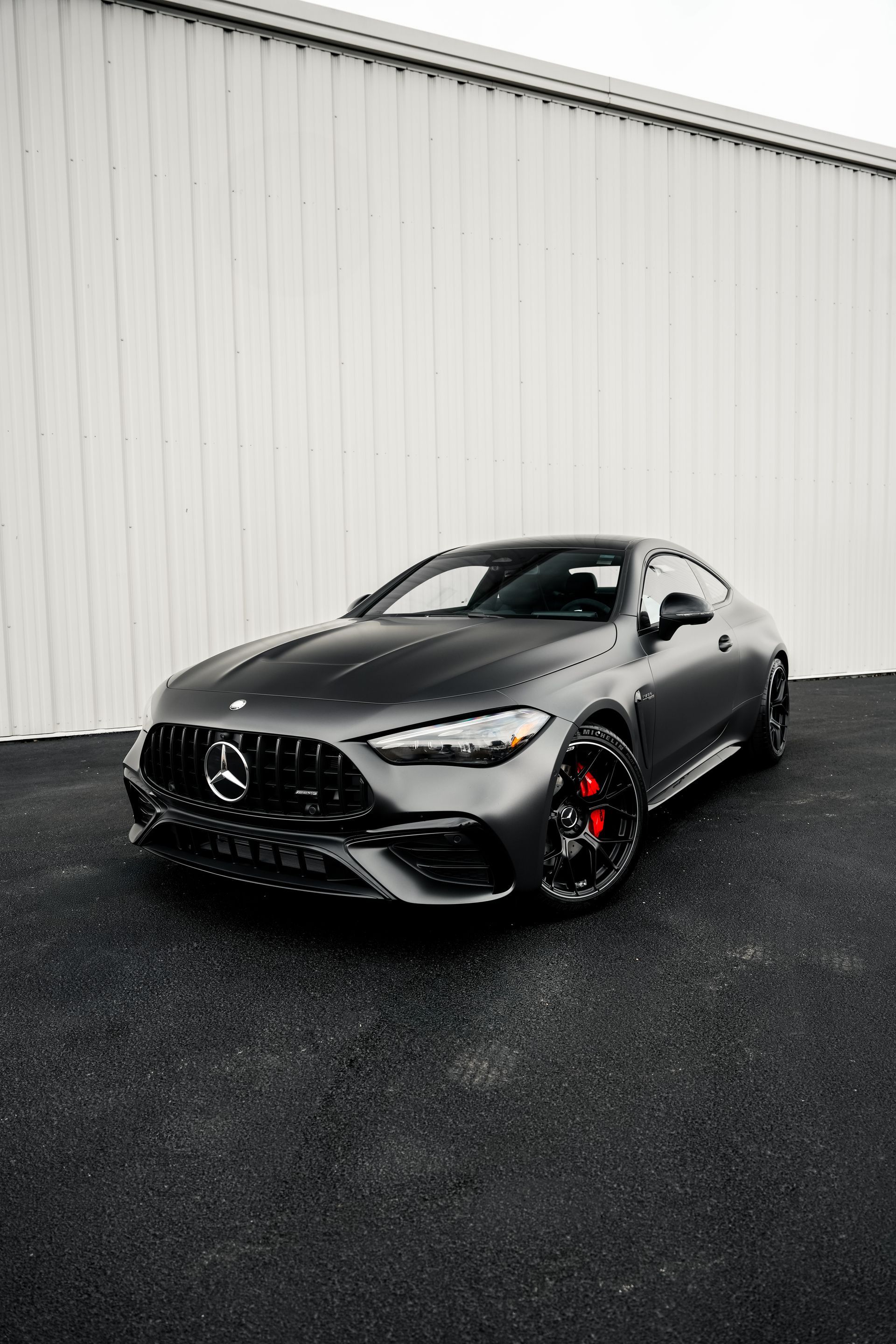 Matte gray sports sedan parked on wet pavement beside a white warehouse wall