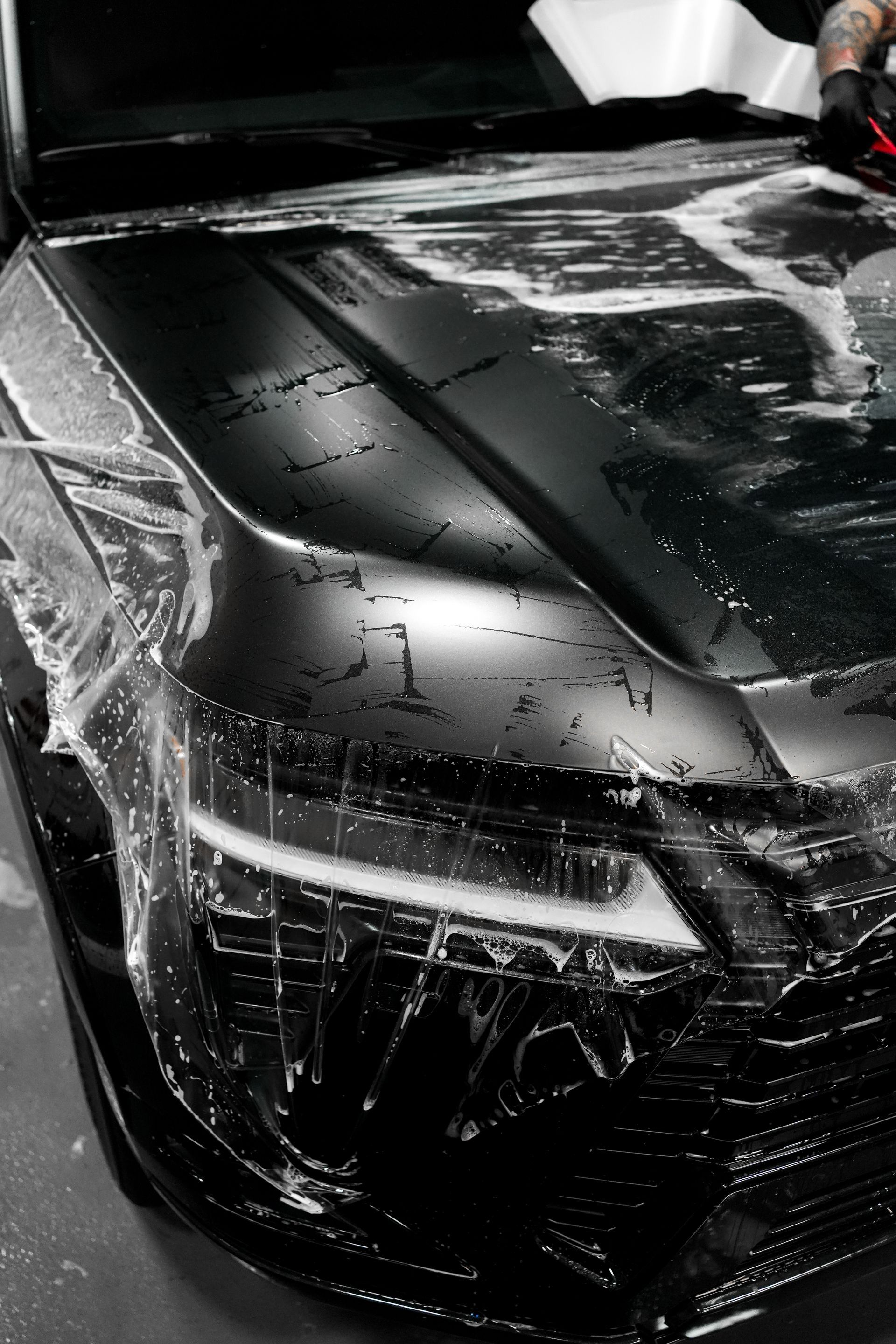 Black sports car being washed, with water and soap suds on the hood and windshield