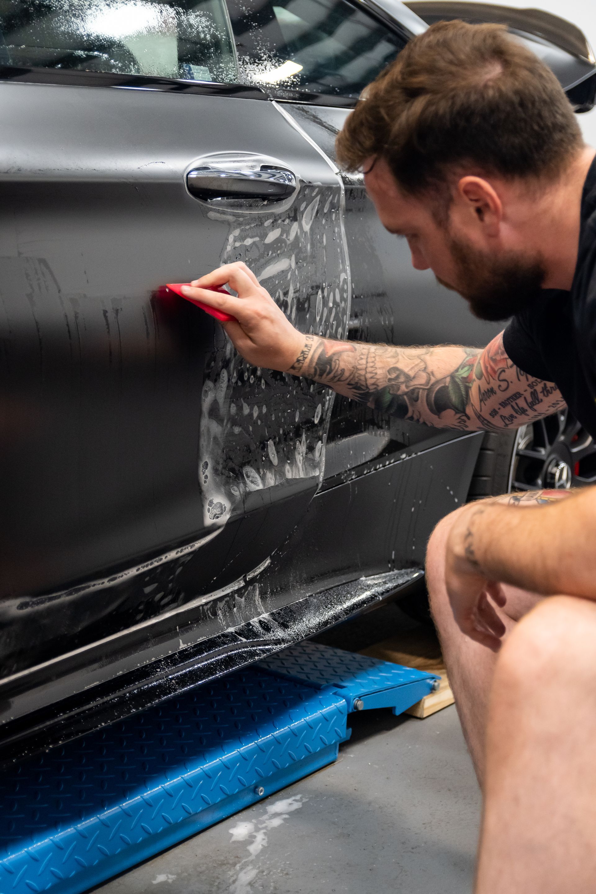 Man scrapes a black car door with a red squeegee while applying window film or wrap.