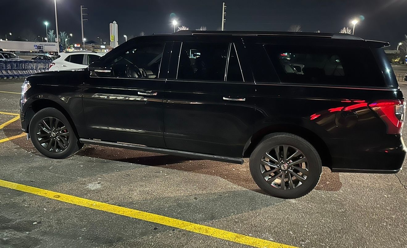 Black SUV parked at night in a lit parking lot, side view with yellow lines on wet pavement.
