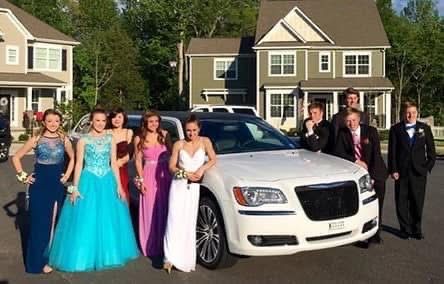 Prom group posing beside a white limousine in a suburban driveway with houses in the background