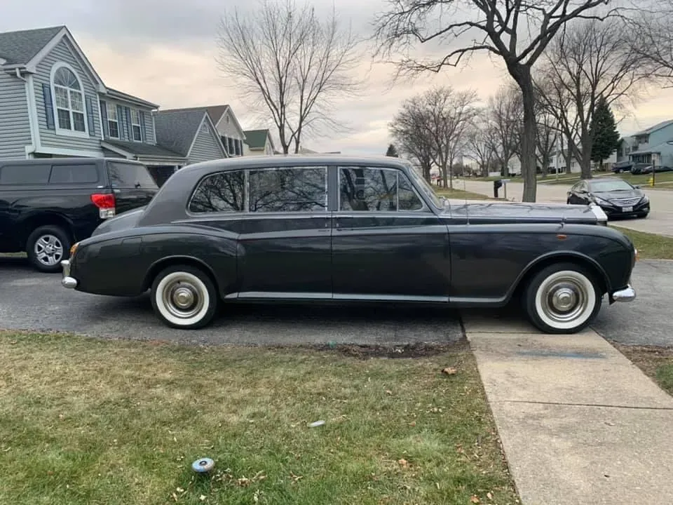 Black vintage sedan parked in a suburban driveway beside a sidewalk, with houses and leafless trees behind it