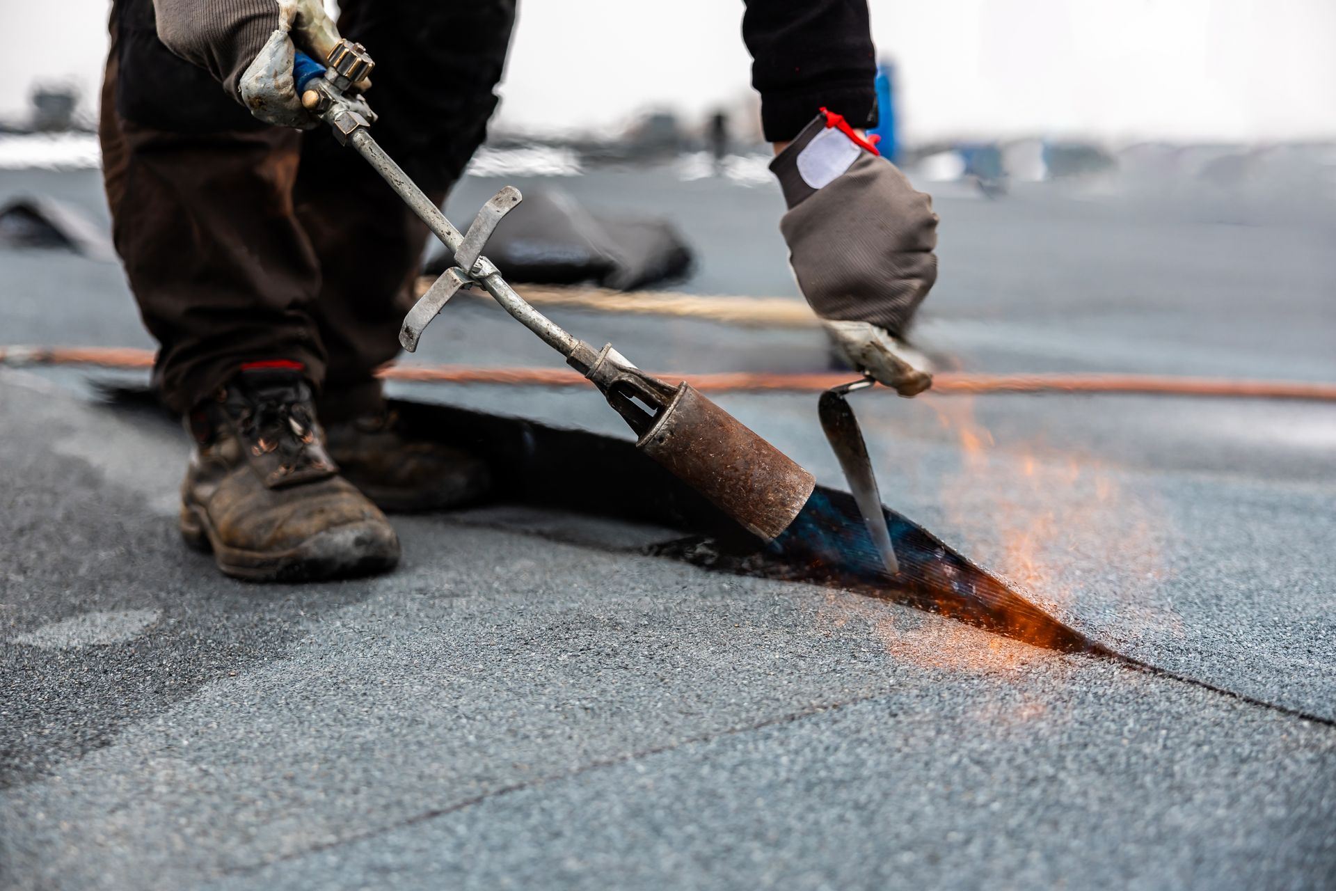 Roofer using a torch to seal seams on a flat, dark gray roof, wearing gloves and work boots.
