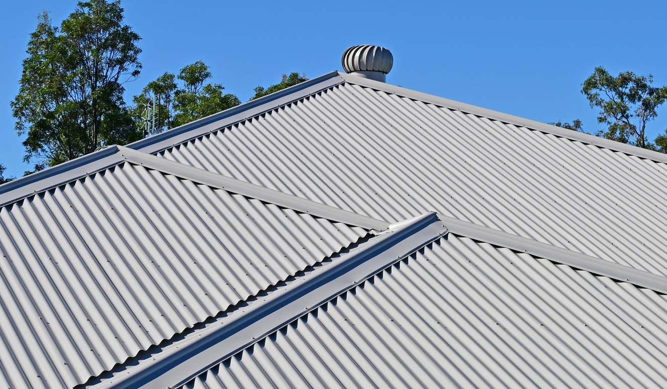Gray corrugated metal roof with a rotating vent and trees against a blue sky.