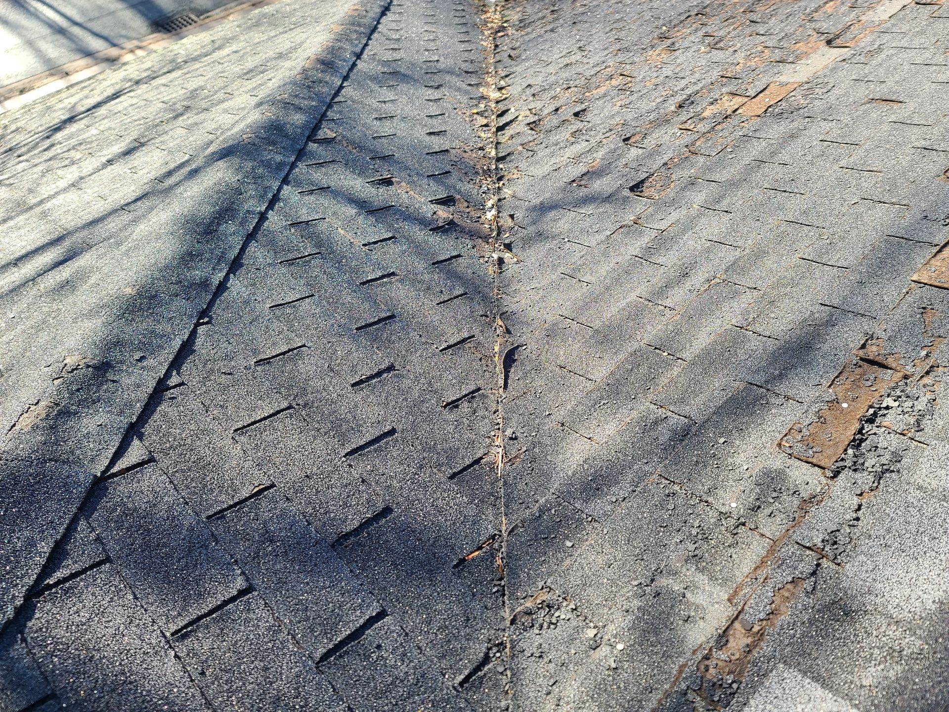 Close-up of a weathered asphalt shingle roof, showing damaged and missing shingles in sunlight.