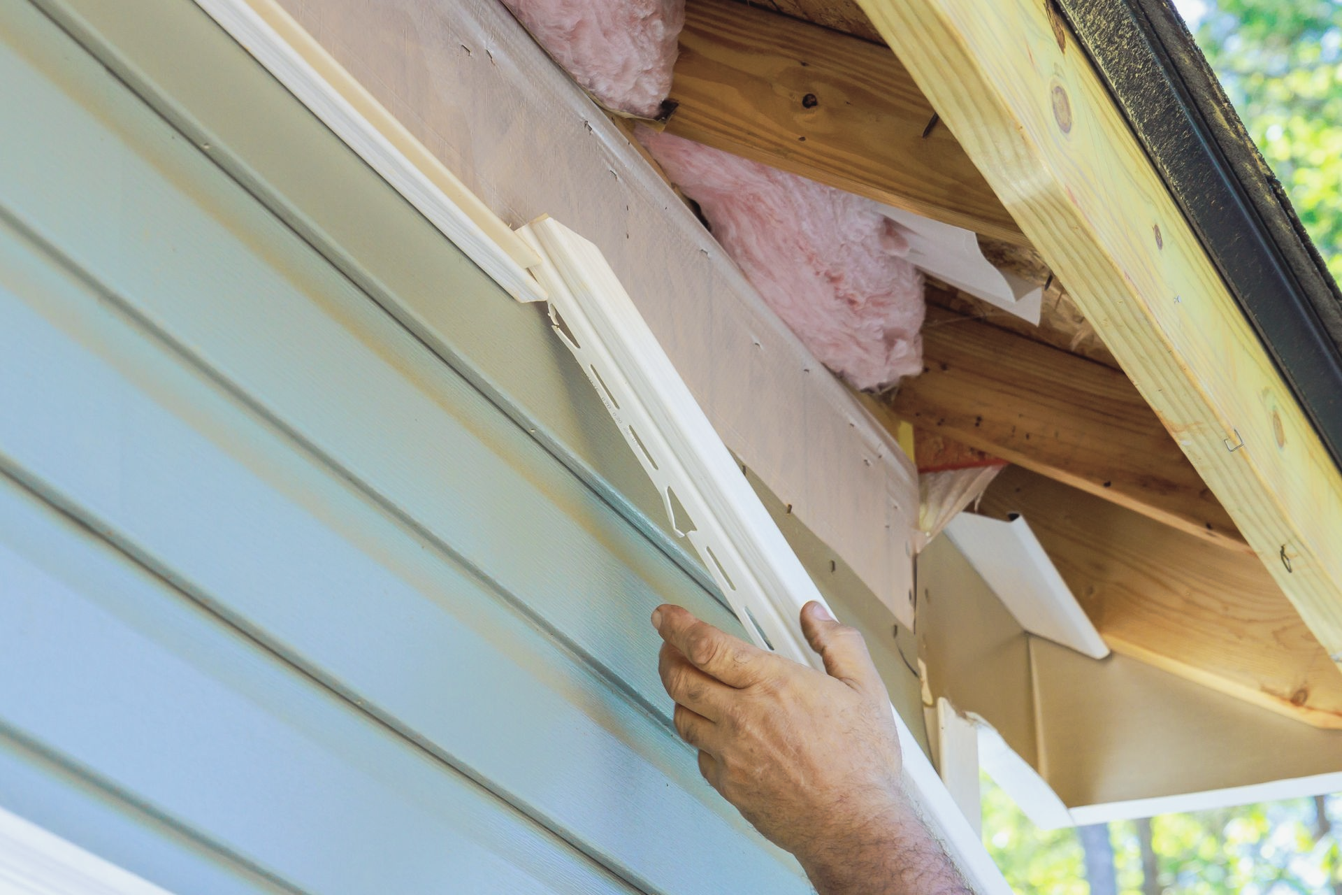 Person installing white siding trim on a light blue house exterior near the roof.