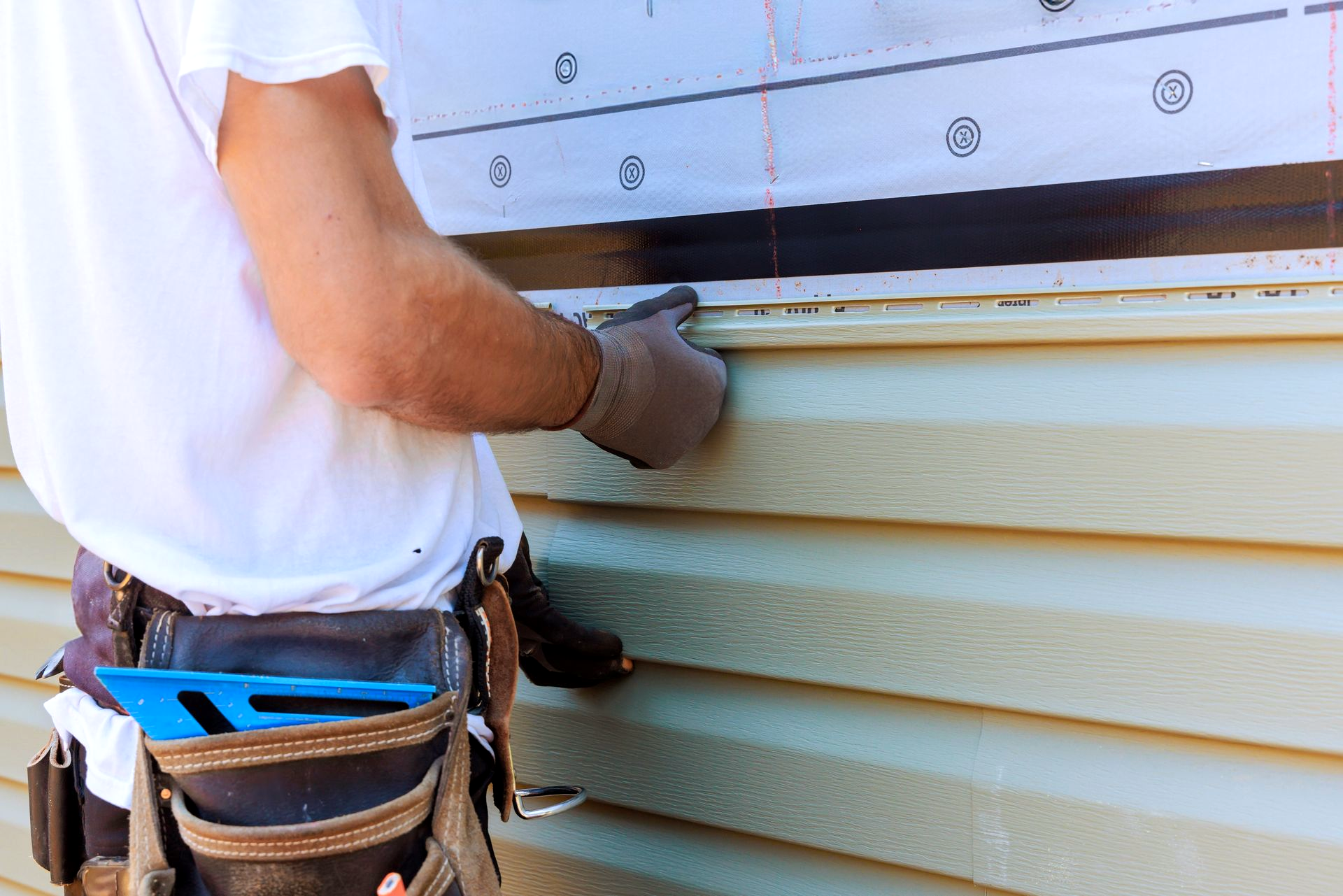 Person installing siding on a building, wearing work gloves and a tool belt.