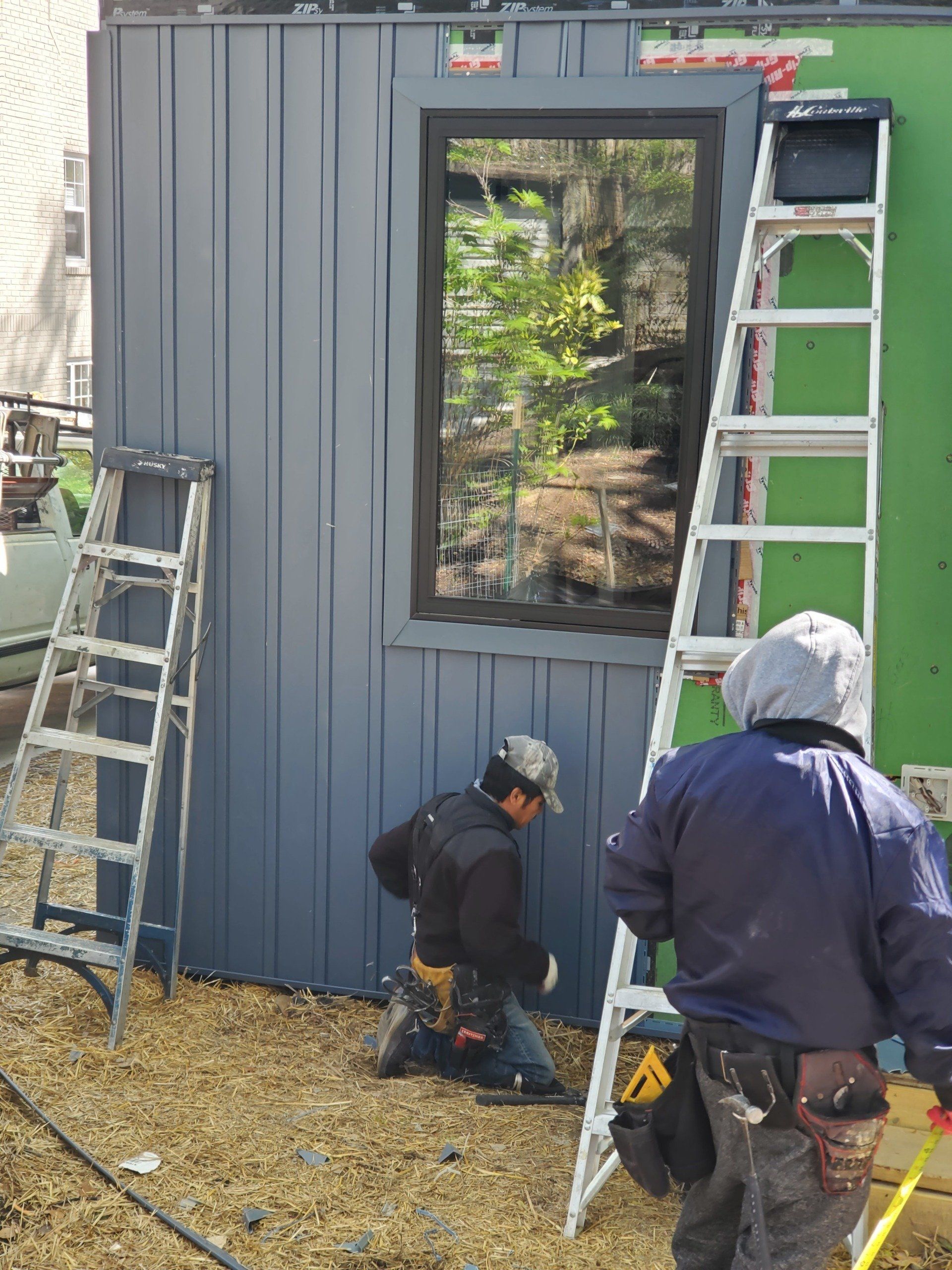 Construction workers installing siding around a window on a building exterior. Ladders and tools are present.