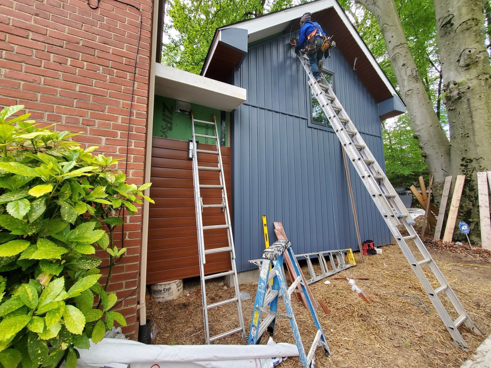 Construction worker on a ladder siding a small building with blue paneling. A brown panel facade is also present.