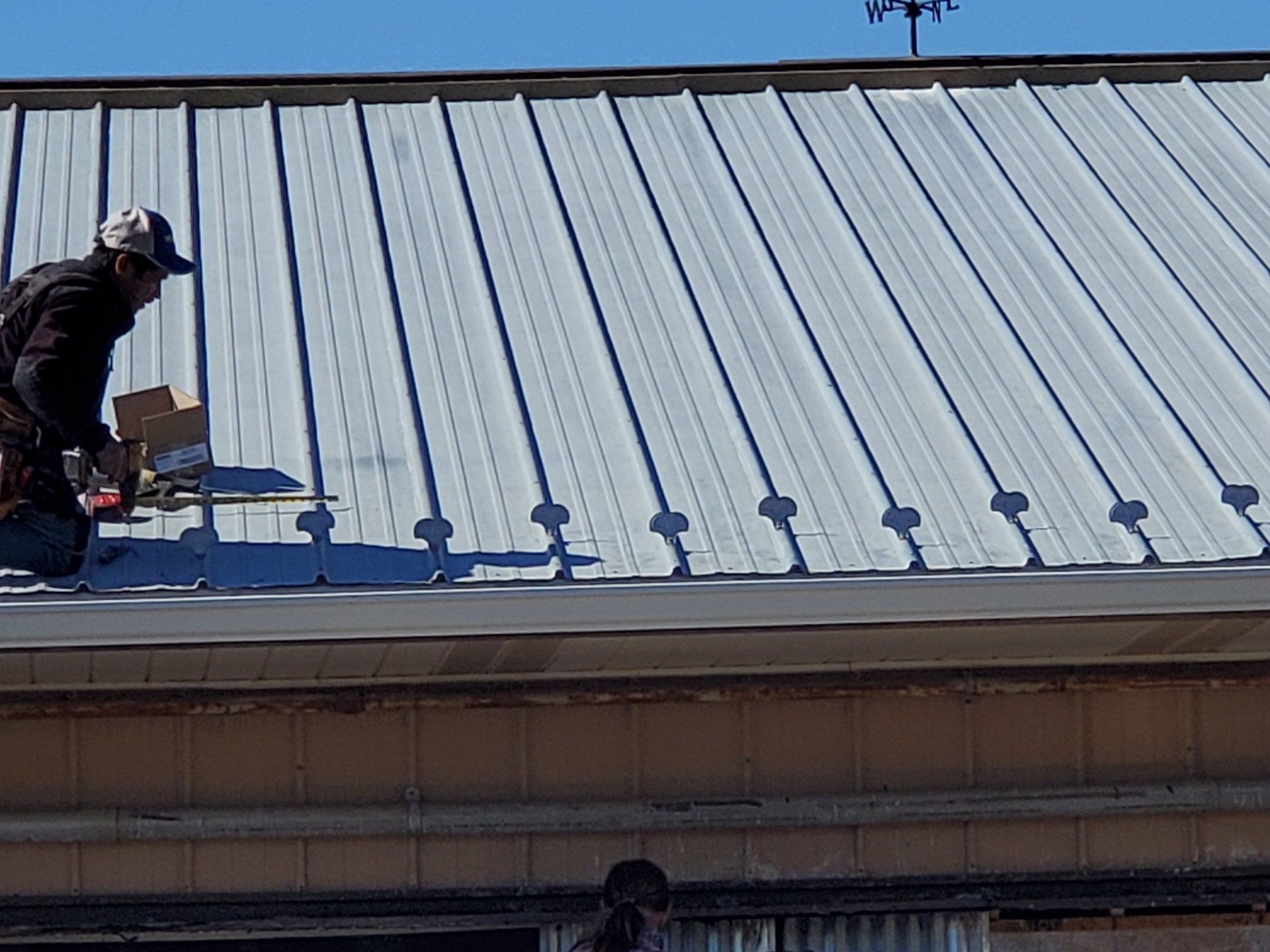 Person on a silver metal roof, working with tools near the edge of the building.