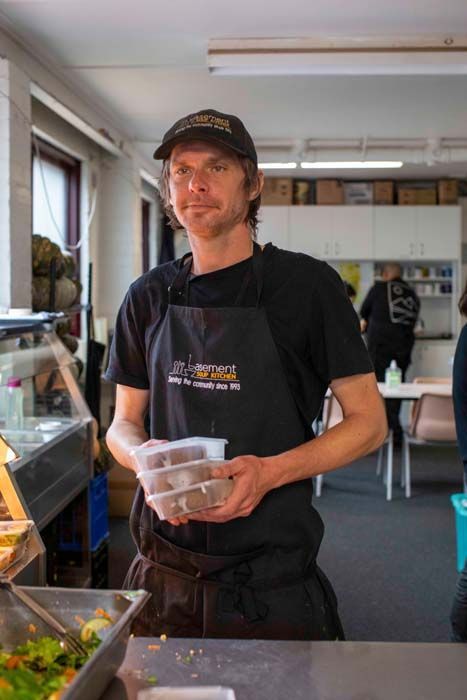 Man in black apron holding a plastic container behind a food counter in a bright kitchen