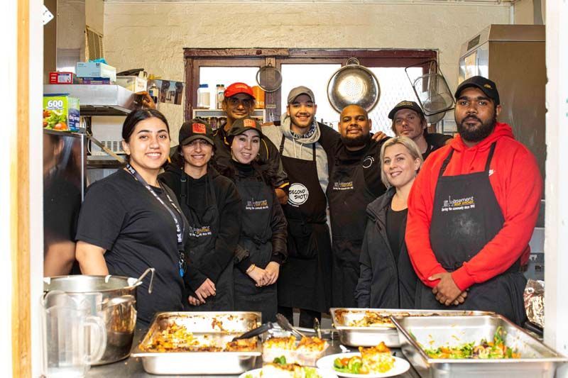 Group of people in a kitchen serving trays of food, smiling at the counter.