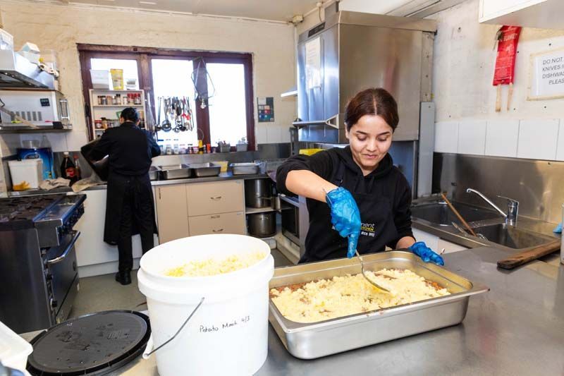 Worker preparing food in a commercial kitchen, filling a tray of baked dish with blue gloves