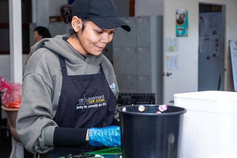 Person in a gray hoodie and black apron preparing food at a counter, with white containers nearby