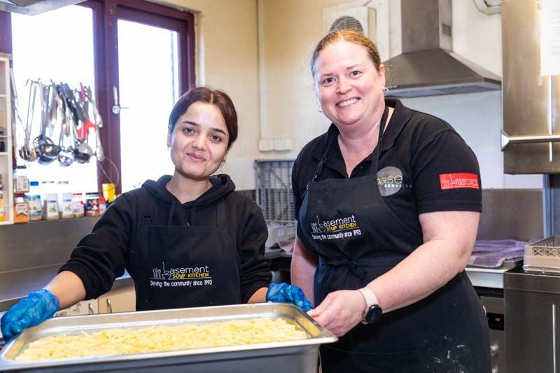 Two kitchen staff in black aprons holding a tray of baked food in a commercial kitchen