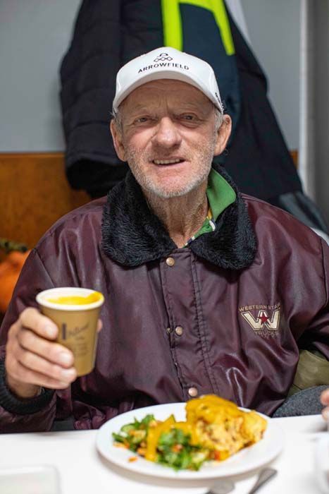 Person in a white cap and maroon jacket holding a drink, seated at a table with a plated meal.