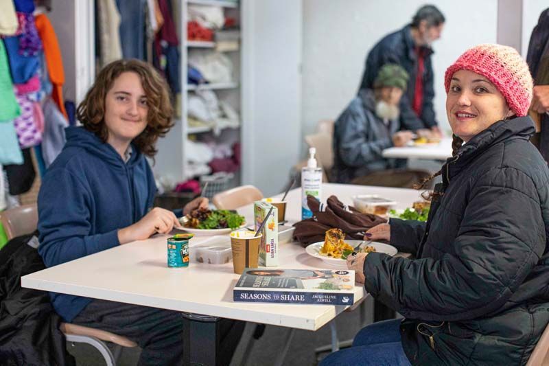 Two people seated at a market table with food and drinks, smiling in a busy indoor setting