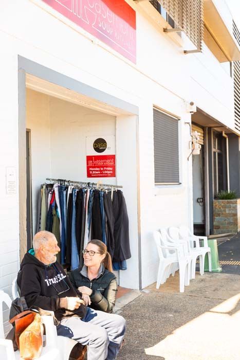 Two people sitting outside a shop entrance beside a clothing rack and white chairs