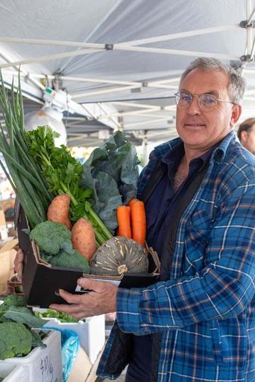 Man holding a box of fresh vegetables at an outdoor market under a white canopy.