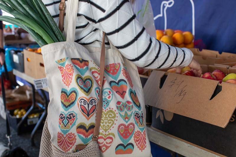 Person reaching for produce at a market, wearing a striped shirt and heart-patterned tote bag